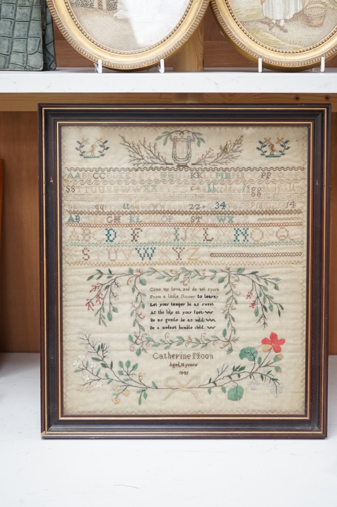 A framed 19th century alphabet sampler by Catherine Noon, dated 1841. Embroidered at the top with a harp and two birds and rows of alphabet and numbers, a central cartouche of verse below and her name and date above a fl